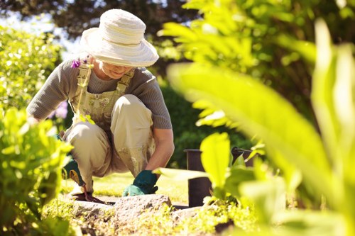 A woman in a garden during daytime, engaged in gardening activities near a flower bed with a variety of plants, including leafy shrubs and flowering borders. She is wearing a large sunhat, glasses, and gardening gloves, and appears to be using a small tool to tend to the garden soil. The garden features a well-maintained lawn with lush green grass, edged by flower beds and mature trees in the background providing shade. Paving stones or stepping stones are visible along the borders of the flower beds, creating a neat walking path. The environment is bright and sunny, suggesting good weather, with sunlight filtering through the foliage, highlighting natural textures and vibrant green tones. This outdoor space demonstrates typical features of a landscaped backyard or front garden in Chingford, suitable for gardening services such as hedge trimming, planting, and lawn care offered by Hedge Trimming Chingford.