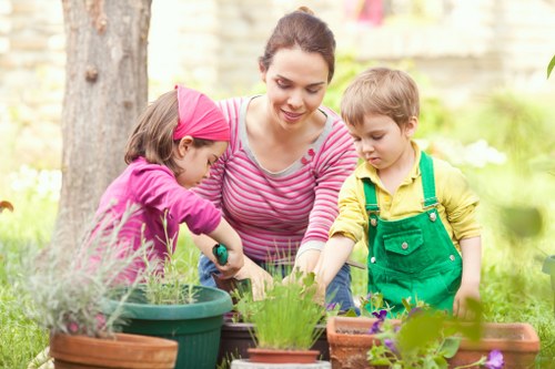 A woman with dark hair tied back, dressed in a pink and white striped long-sleeve top, is engaging with two young children in a garden setting. One child, wearing a pink headscarf and purple long-sleeve top, is tending to potted plants, while the other child, dressed in a yellow shirt and green overalls, is observing nearby. The garden features a variety of potted plants placed on a wooden surface, with a large tree trunk and a brick wall in the background. The scene is illuminated by natural daylight, suggesting a clear, sunny day, and the garden area includes lush green grass and blooming plants, reflecting well-maintained landscaping typical of outdoor garden spaces in Chingford. This outdoor environment exemplifies a family-friendly garden dedicated to plant care and outdoor activities, aligning with gardening services like those offered by Hedge Trimming Chingford.