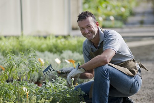 A man with short dark hair, wearing a light grey shirt, dark blue jeans, and a brown gardening apron, is crouching in a well-maintained garden during daylight hours. He is smiling and holding a small garden rake, actively tending to a flower bed. The garden features a neatly trimmed hedge in the background, a lush green lawn in the foreground, and a variety of low-growing plants and flowers with green foliage and hints of yellow and orange blossoms. The ground surface around the flower beds appears to be soil or mulch, and a wooden fence is visible in the background, suggesting a private outdoor space within Chingford. The natural lighting and outdoor environment indicate a pleasant, mild weather condition. This scene exemplifies professional gardening and hedge trimming services aimed at maintaining a tidy and attractive garden landscape, as part of the offerings from Hedge Trimming Chingford in London postcode E4.